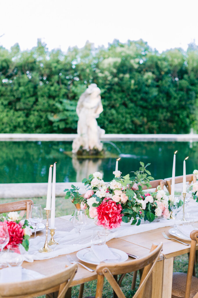 Une table en bois décoré avec des bouquets de fleurs rouges et des bougies blanches au bord d'un étang. 